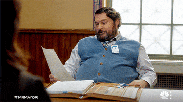 TV gif. Bobby Moynihan as Jayden Kwapis in Mr Mayor examines a sheet of paper while leaning back in his chair and then falling backwards with his feet in the air.