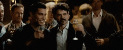 Video gif. Two men stand around with other men who all hold different glasses and bottles of alcohol. They all lift their glasses and bottles up in celebration as one of the men says, “Cheers.” The two men lift their glasses up and link them together. 