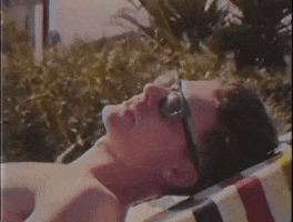 Video gif. Man wearing sunglasses lies on a beach chair. Text slides in from both sides, "Amazing. What the heck."