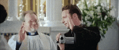 Movie gif. Andrew Lincoln as Mark in Love Actually high fives a priest while holding a small video camera.