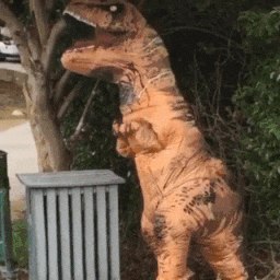 Video gif. A person wearing an inflatable T-Rex costume is standing in front of a bush and eagerly waves with their short, stubby paw. 
