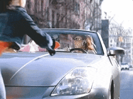 Music video gif. From the video for Stupid Girls by Pink, the singer drives a convertible with various devices in hand, running over a pedestrian who hits her windshield, as she looks shocked.