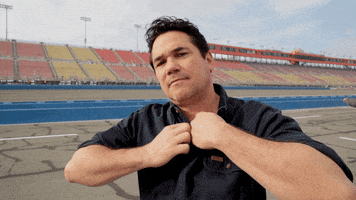 Celebrity gif. Dean Cain stands on a race car track. He looks at us and rips over his button shirt to show his black shirt. When he rips his shirt open he says, “Boom!”