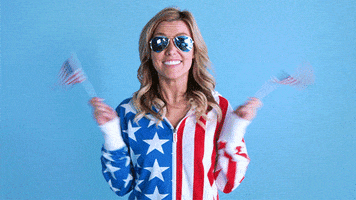 Video gif. A woman wearing blue aviators is wearing an American flag printed shirt and grins happily as she holds two small American flags and waves them around.