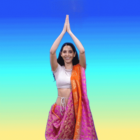Video gif. Woman wearing a magenta, orange, and gold sari stands with her palms pressed together above her head in front of a background that fades from blue to yellow. As she opens her arms out to the side, a rainbow expands between them, raining flowers while two birds fly off in opposite directions. Text, "It's spring!"