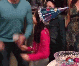 Video gif. Young girl in a pink turtleneck and a sparkling birthday cone hat is overcome with excitement at what appears to be a family birthday. A man yells something towards us behind her and, all of a sudden, she picks up a bowl of colorful balls and smashes it on the floor in front of her, raising her arms in the air like she's totally pumped about it. 