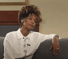 Movie gif. We slowly zoom in on Whitney Houston, who sits on a couch wearing a buttoned-up white shirt and a black headband, as she silently listens to someone offscreen.
