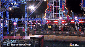 Reality TV gif. Man on American Ninja Warrior swings down and hits the button, causing fireworks to go off around him. He jumps up and screams in excitement.