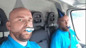 Video gif. In the backseat of a truck, two bald men with similar beards, bright blue shirts, and party hats blow into birthday noisemakers and stare directly at us.